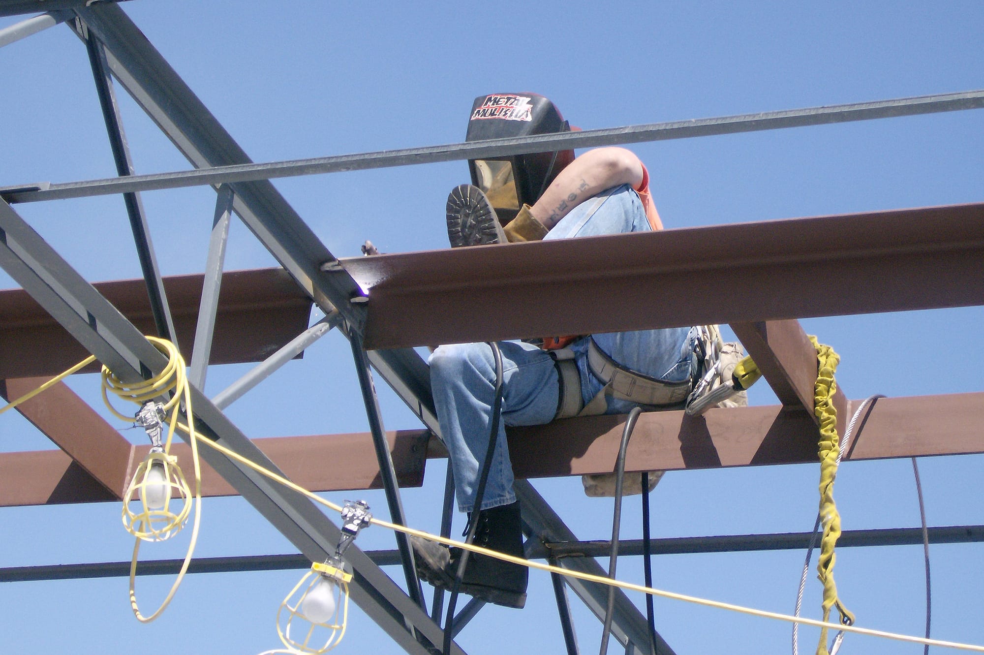 A Vancouver, Wa steel fabricator sits atop high metal beams in a safety harness with a safety mask on for welding.